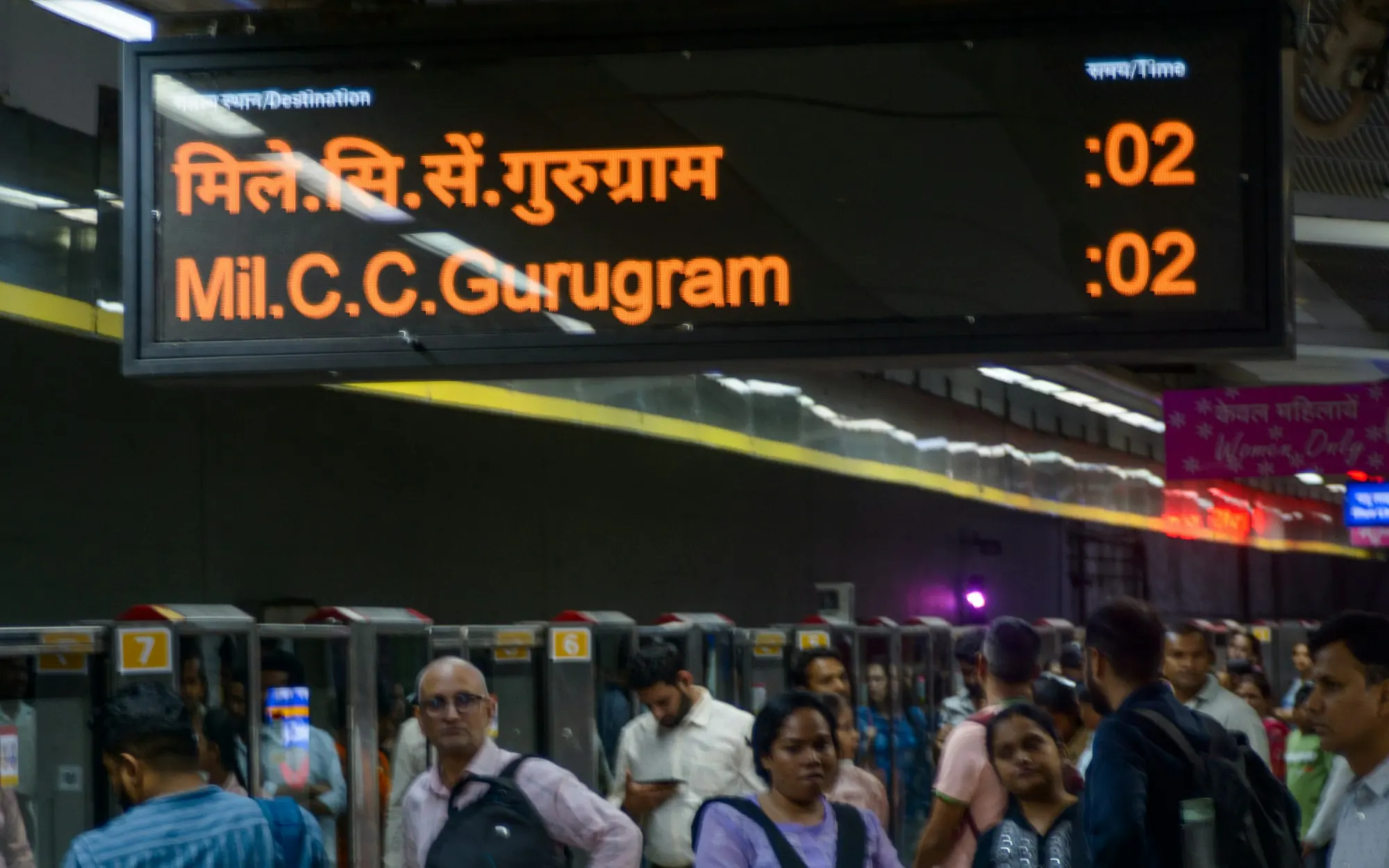 Commuters on a Delhi Metro platform under a departure board: next train to Millennium City Centre Gurugram in two minutes