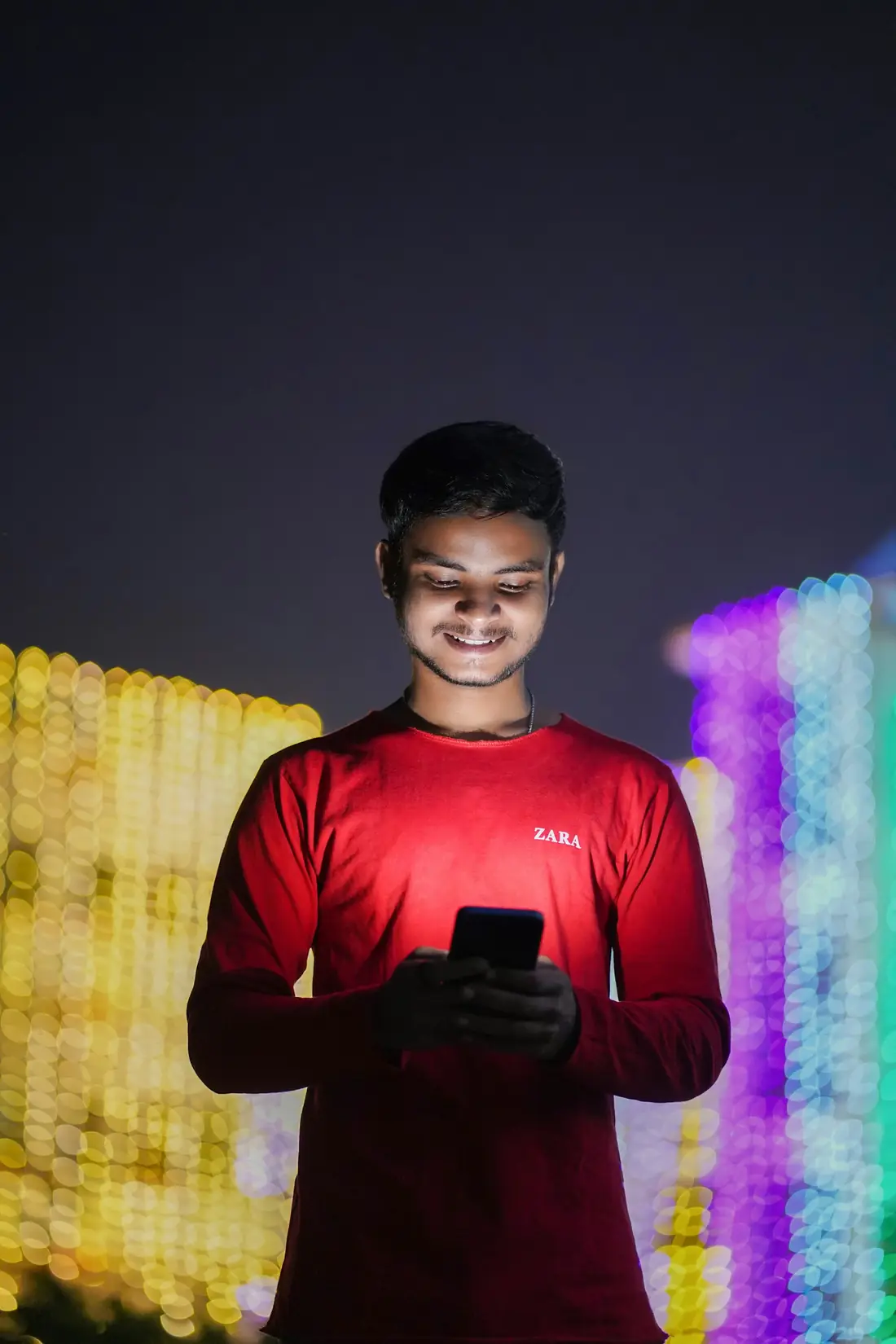 A young man at night, his face lit by his phone screen, with festival lights behind him